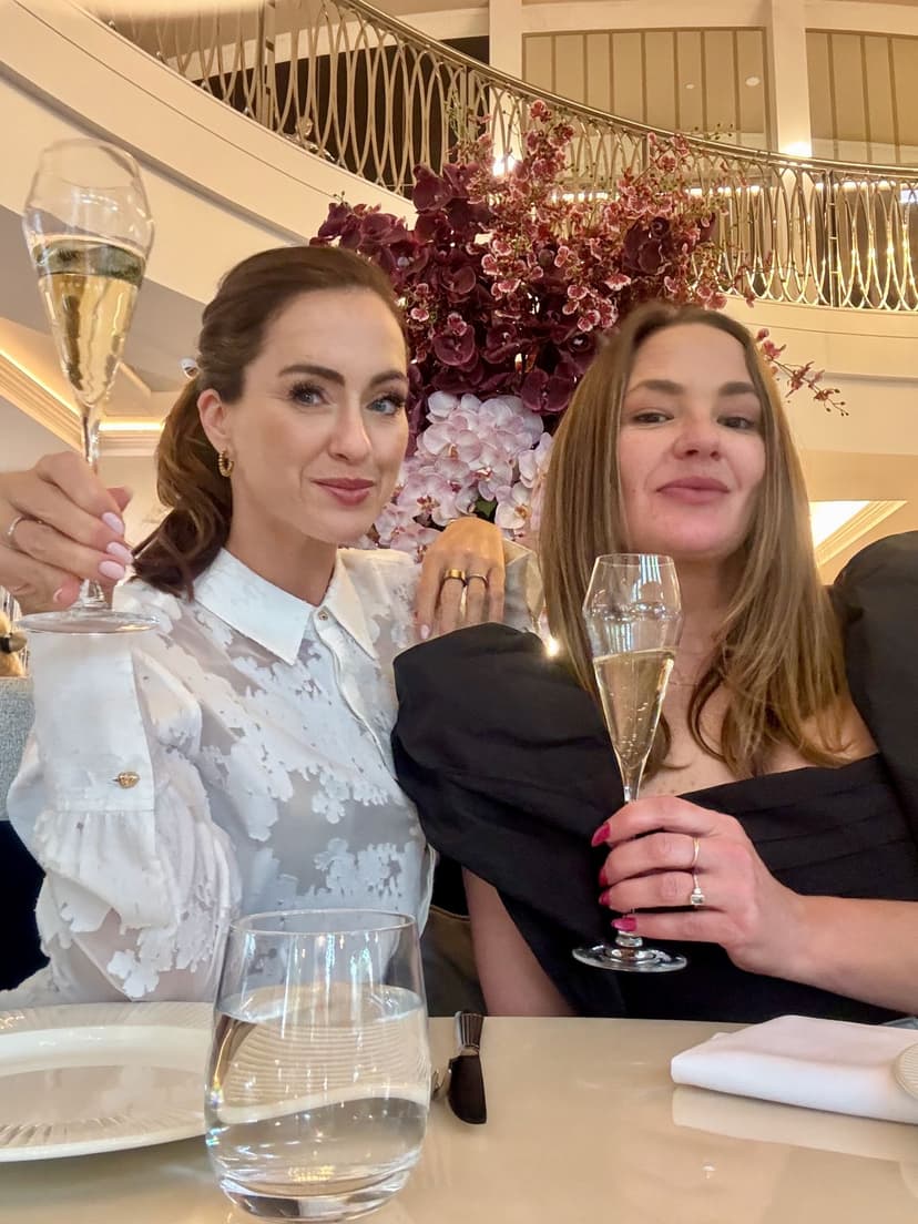 Sigourney and Jayde toasting champagne flutes at the Showroom Bar, The Royce Hotel Melbourne, orchids and the spiral staircase behind them