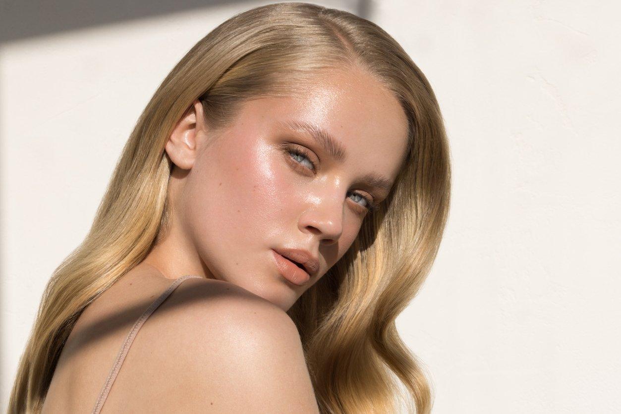 Close-up portrait of a young woman with long, sleek blonde hair and glowing, dewy skin. She gazes intensely at the camera with blue eyes, her face softly illuminated by natural sunlight against a minimalistic white background.