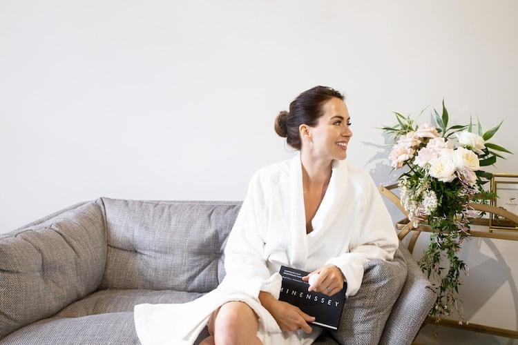 Woman with clay face mask applied, resting on massage bed with flowers and skincare products nearby