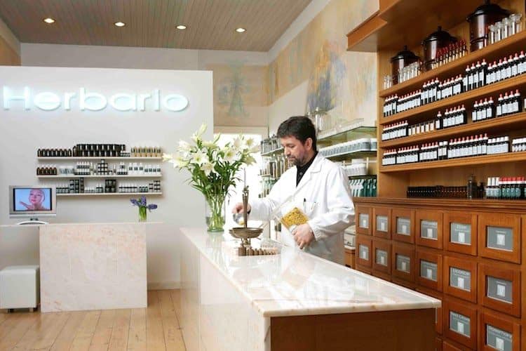 Valli Shubere in white coat at Herbario clinic with shelves of herbal remedies and botanical products