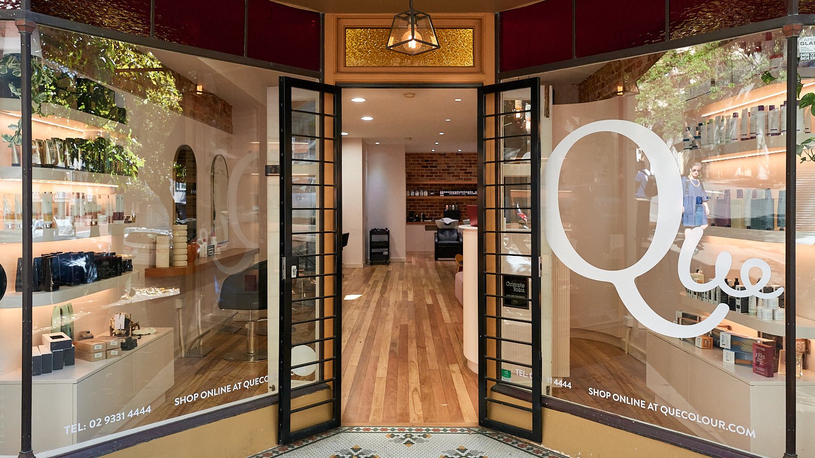 Open doors of the QUE Colour shopfront in Darlinghurst, looking down a timber-floored salon to an exposed-brick wall