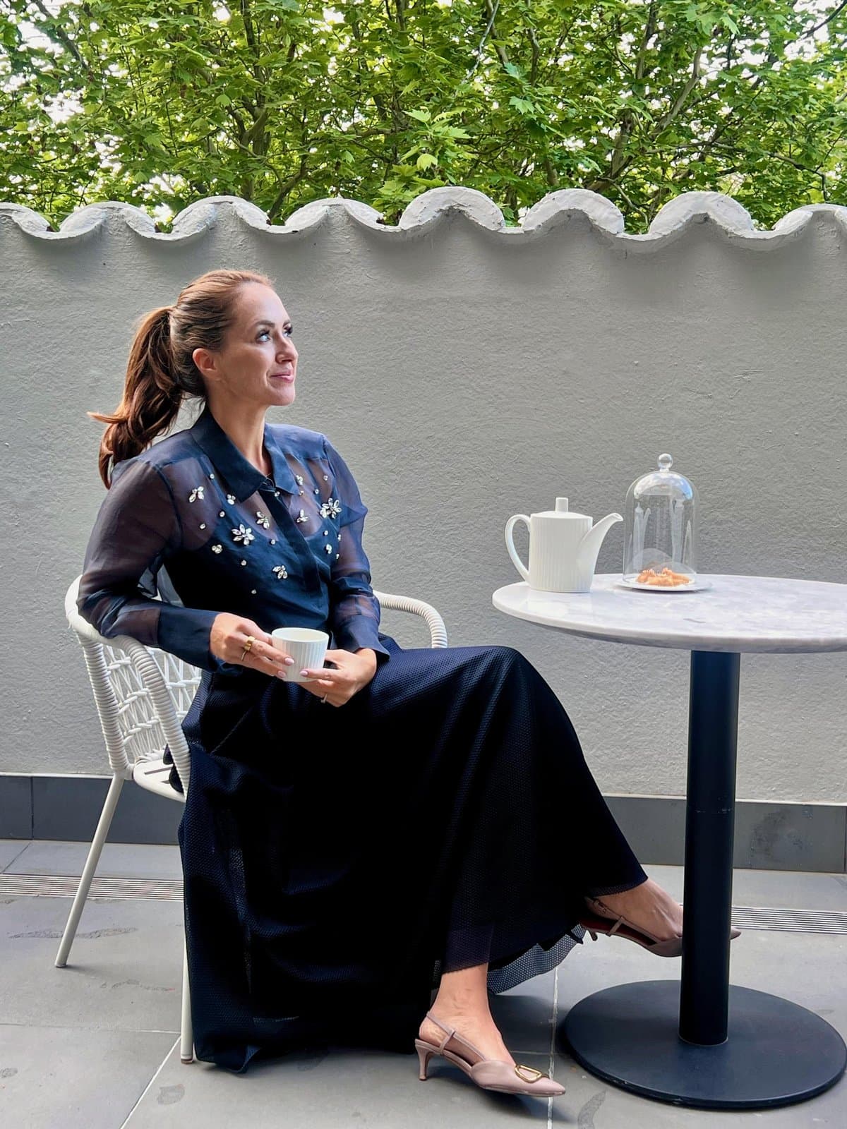 Sigourney seated on the terrace at The Royce Hotel Melbourne, tea in hand against the scalloped garden wall