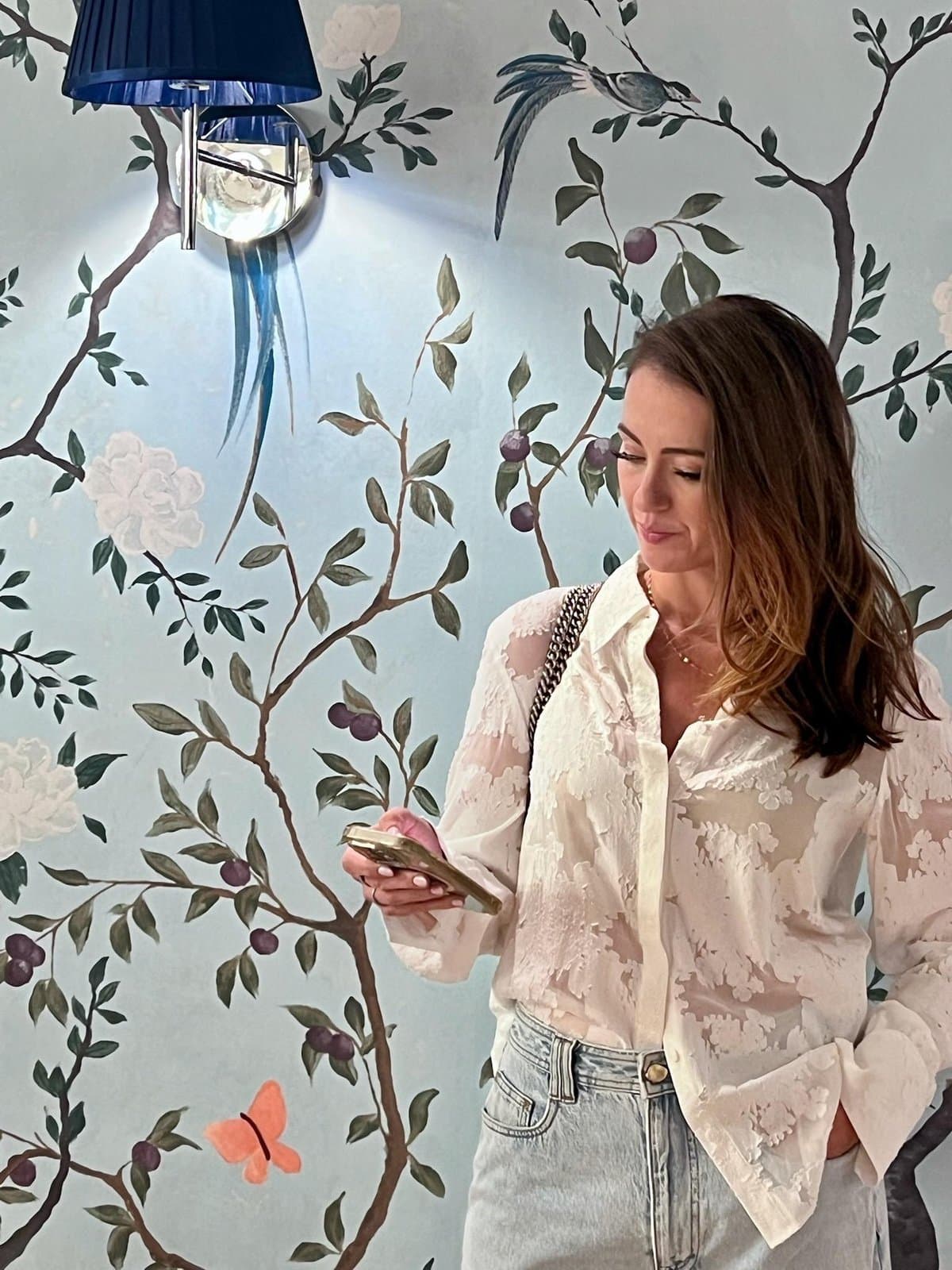 Sigourney checking her phone in a corridor at The Royce Hotel Melbourne, hand-painted chinoiserie wallpaper behind her