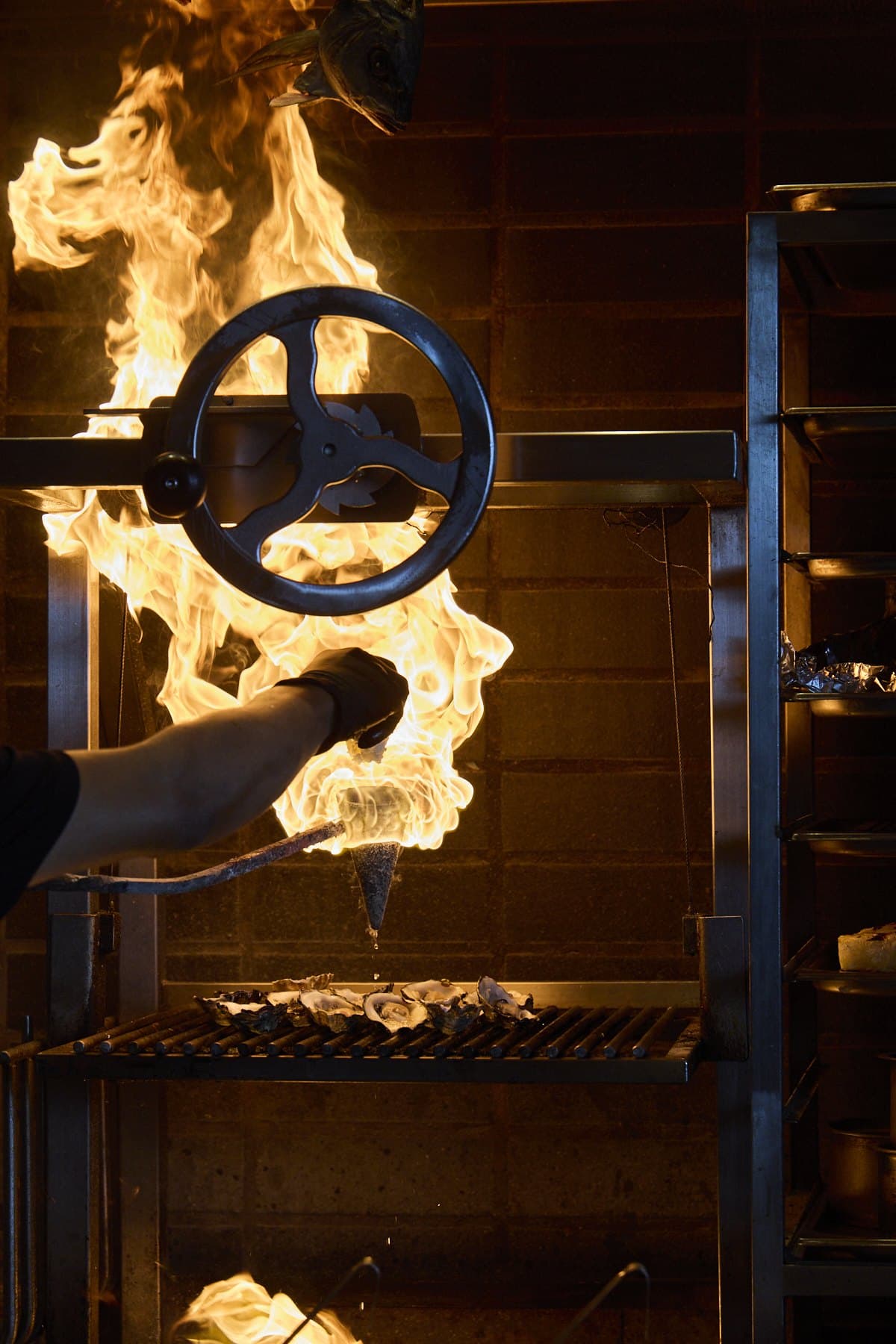 A hand holding a flaming flambadou over the charcoal grill at Lua, flames rising behind the grill wheel