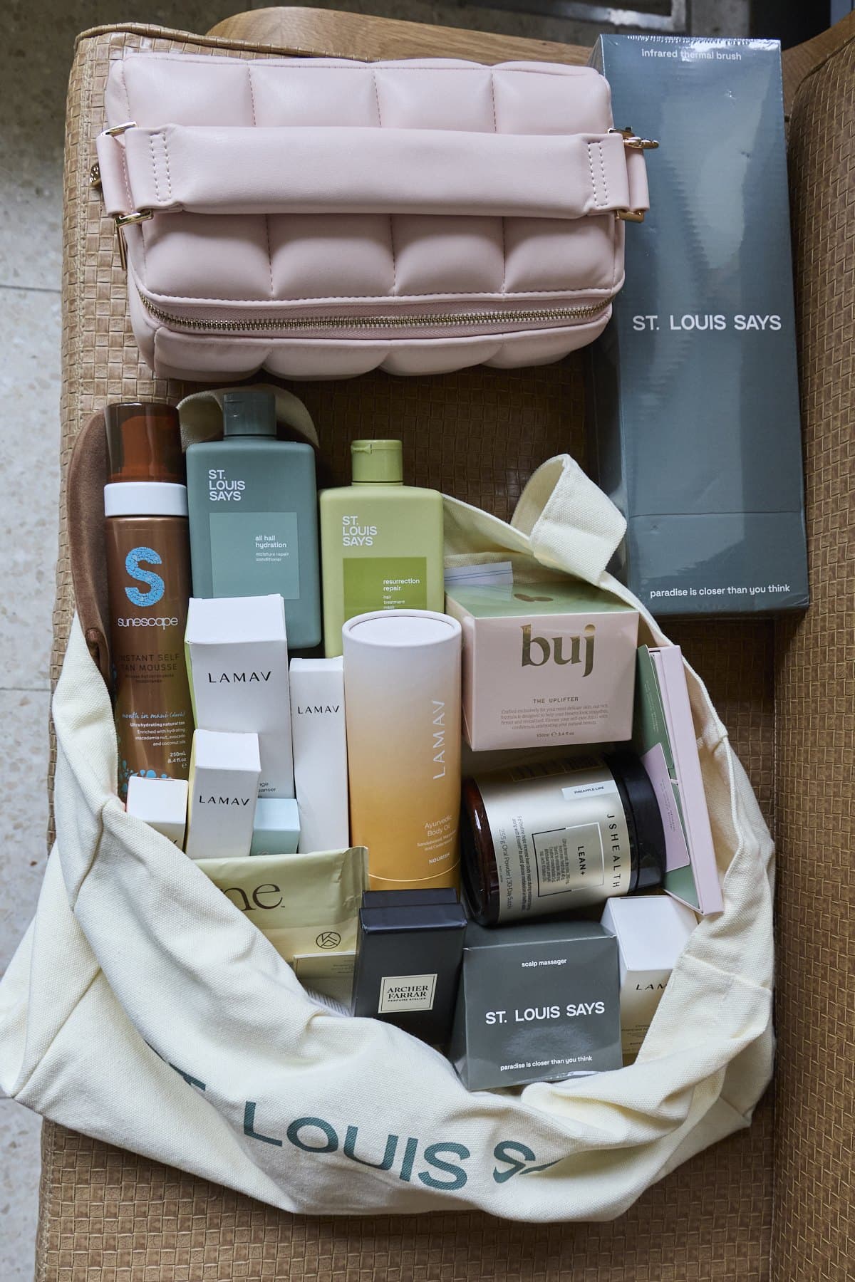A St. Louis Says tote spilling gift products including LAMAV, buj and hair care, beside a pink quilted makeup bag