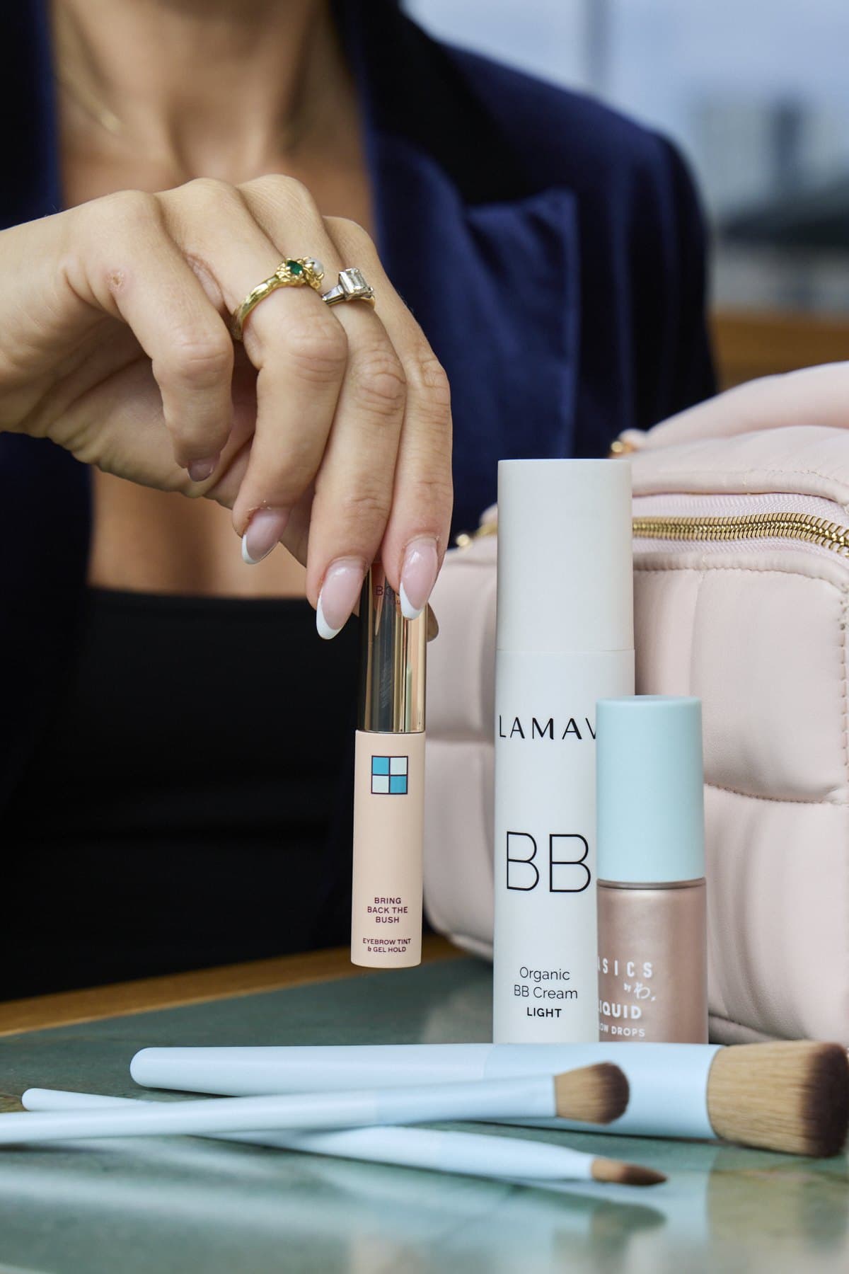 A hand holding a LAMAV concealer above a pink Estetika bag, with a BB cream beside it
