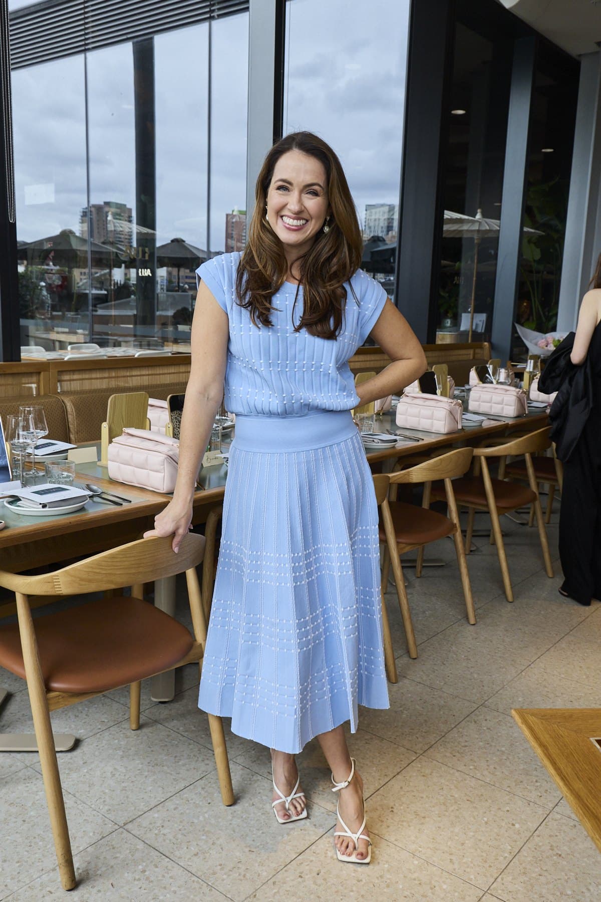 Sigourney in a blue knit dress standing beside the long table at Lua, with harbour windows behind