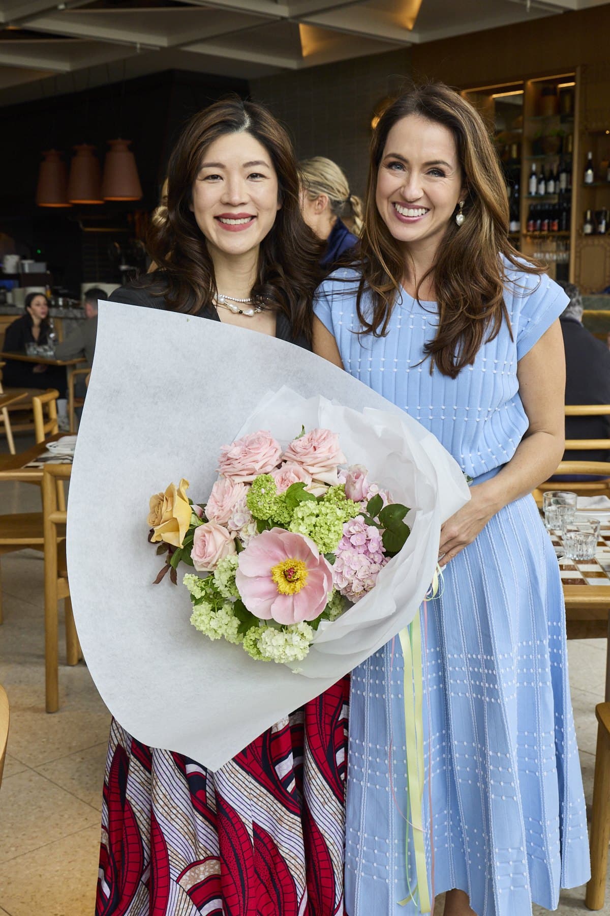 Sigourney in her blue dress holding a bouquet beside a guest in a patterned skirt at Lua