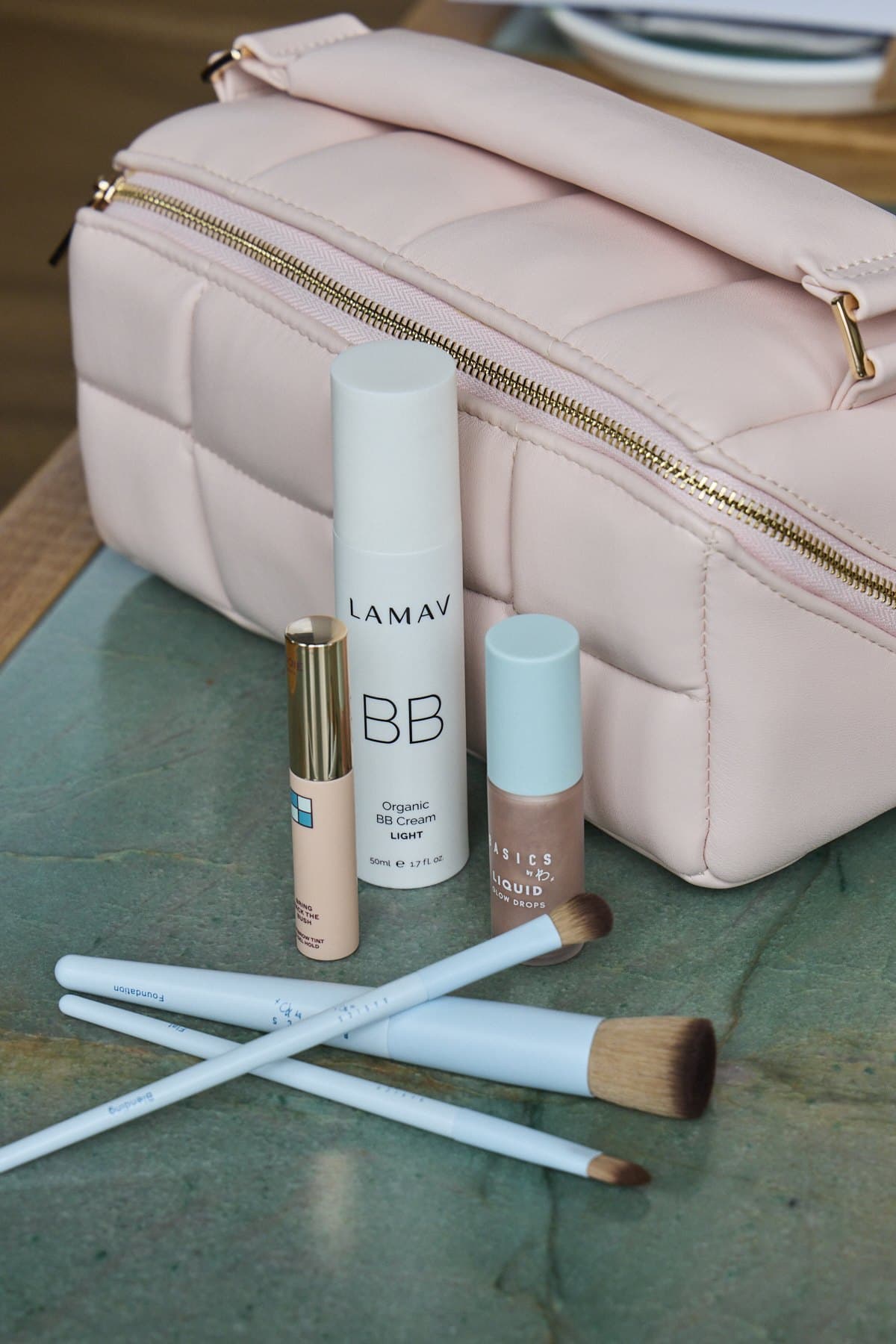 A LAMAV BB cream, a concealer and makeup brushes laid out in front of a pink quilted bag