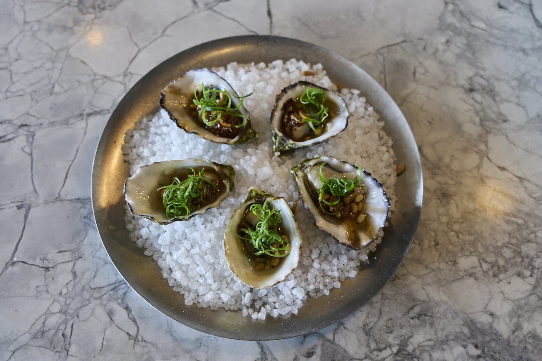 Freshly shucked oysters topped with herbs on a bed of ice in a metal dish
