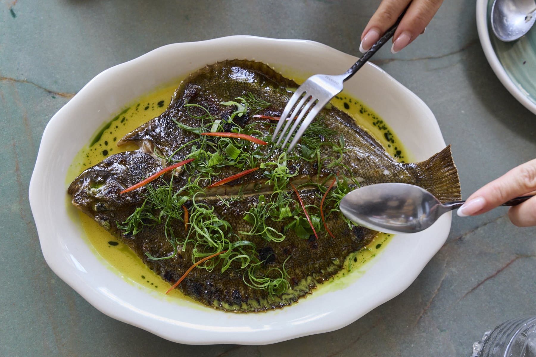 A whole fish in a bright herb and oil sauce in a white dish, being served with a fork and spoon