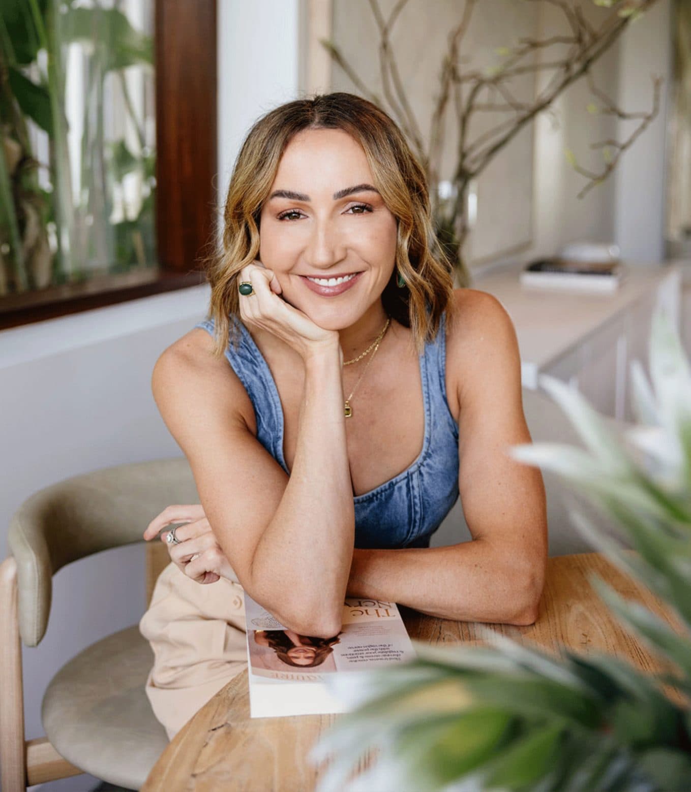 Jessica Maguire smiling at a wooden table in a plant-filled garden room, for Beautiful Inside by Beauticate