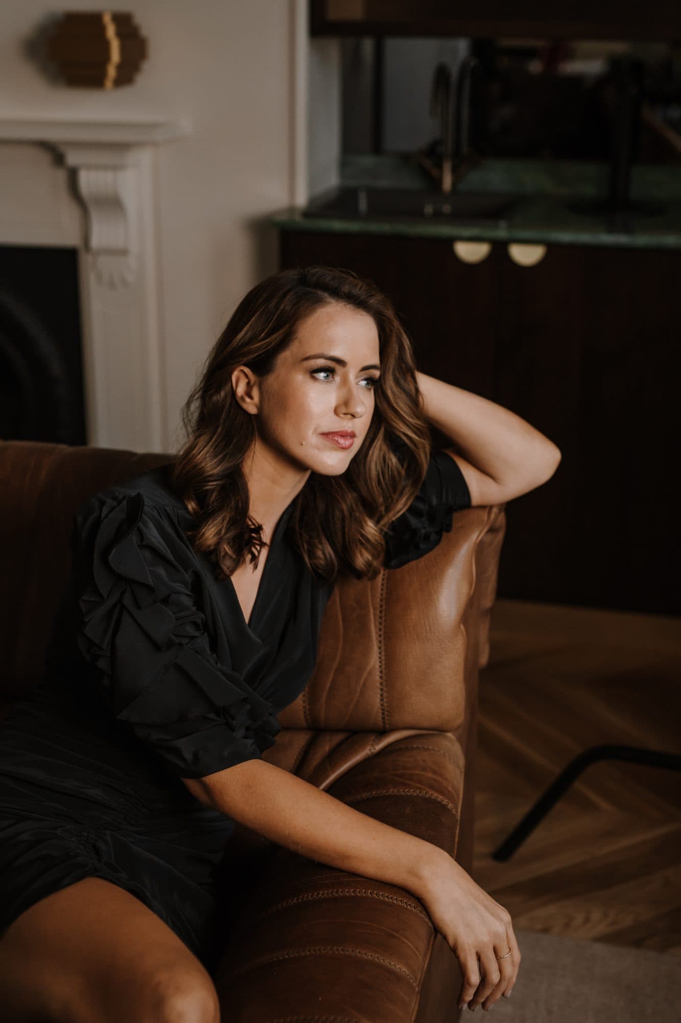 Sigourney Cantelo in a black dress on a leather sofa, looking to the window, for Beautiful Inside by Beauticate