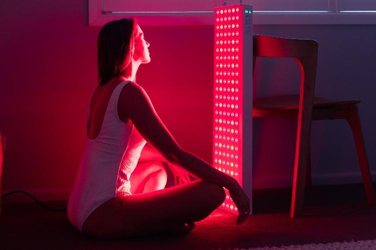 Woman sitting beside an Infraredi red light therapy panel at home, embracing wellness and skin rejuvenation in a calm, minimalist setting.