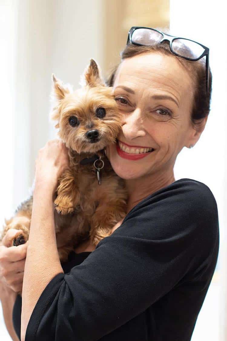Rose-Marie Swift smiling and cuddling a small Yorkshire Terrier, in a black top with glasses on her head