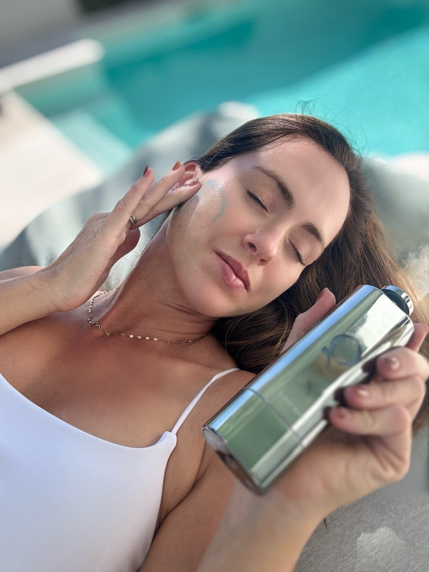 Sigourney Cantelo lying poolside with a streak of blue serum on her cheek, holding the Blue+ Face Serum