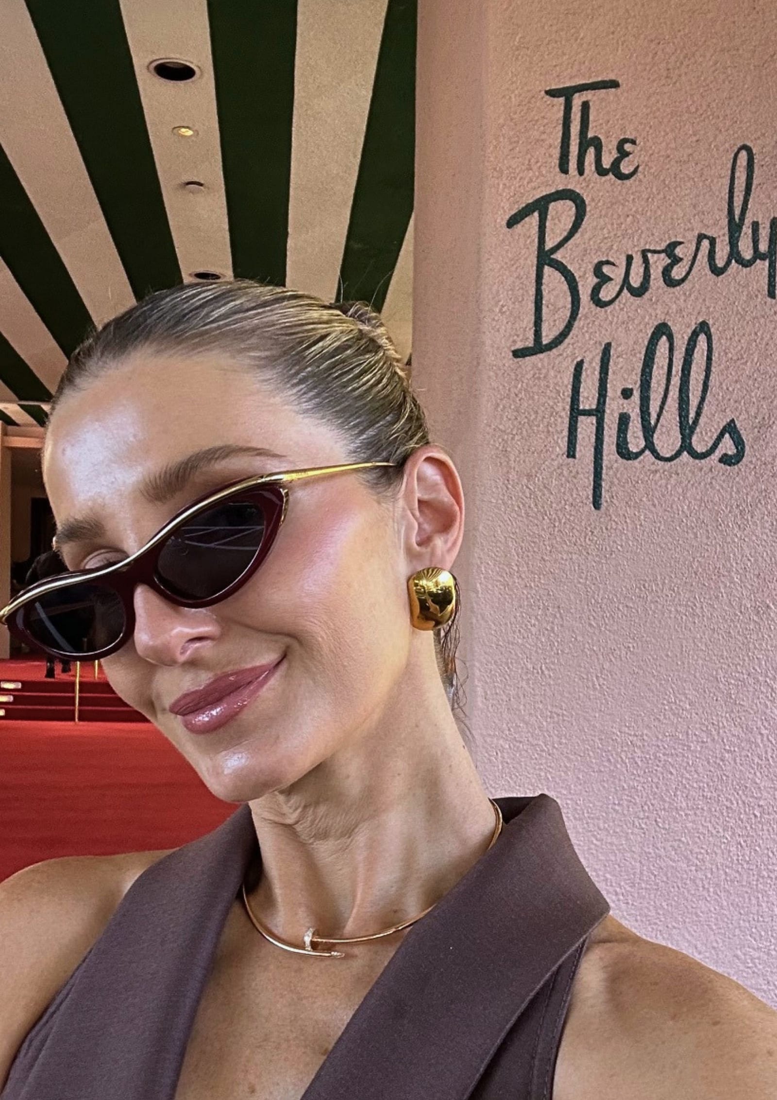 Kate Waterhouse taking a selfie outside The Beverly Hills Hotel beside the iconic pink wall with green script lettering, wearing tortoiseshell cat-eye sunglasses and gold statement earrings