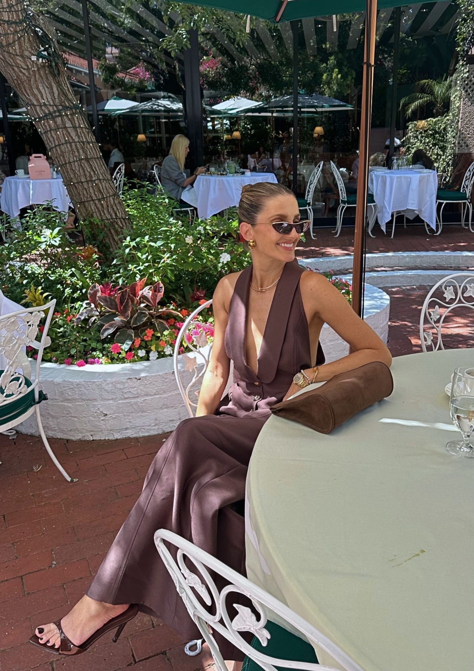 Kate Waterhouse sitting at a garden terrace table at The Beverly Hills Hotel, wearing a chocolate brown halterneck and wide-leg trousers, gold jewellery and cat-eye sunglasses