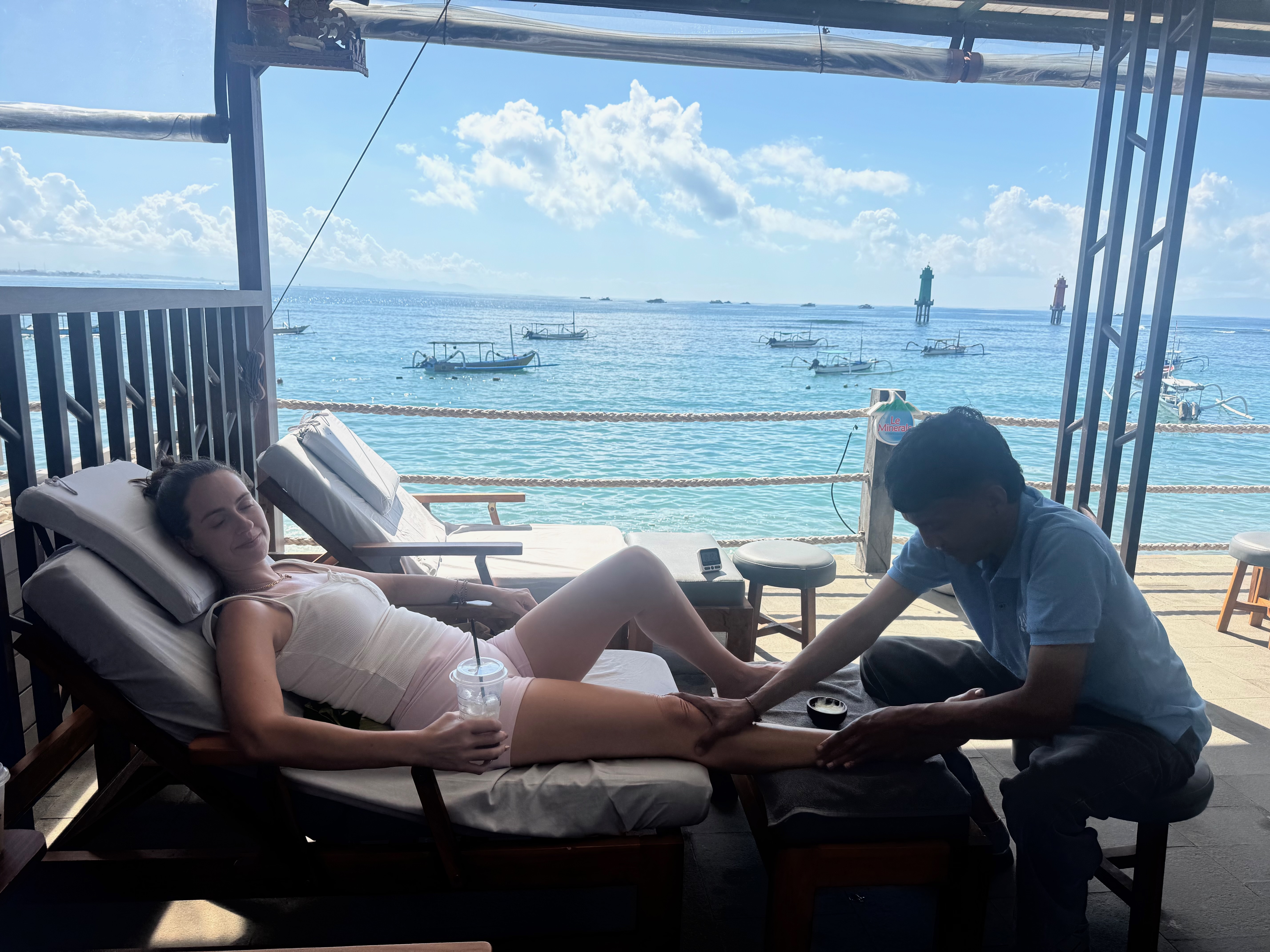 A family getting foot massages at an outdoor spot near the Sanur port, with the harbour in the background