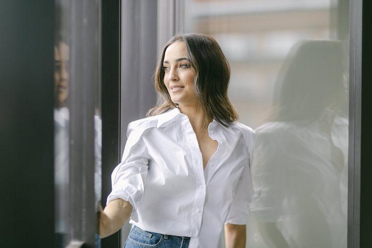 Abbey Gelmi in a white shirt and jeans leaning between glass doors, smiling, city backdrop