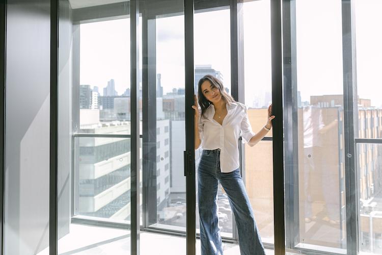 Abbey Gelmi in floor-to-ceiling glass doors wearing a white shirt and wide-leg jeans, city skyline