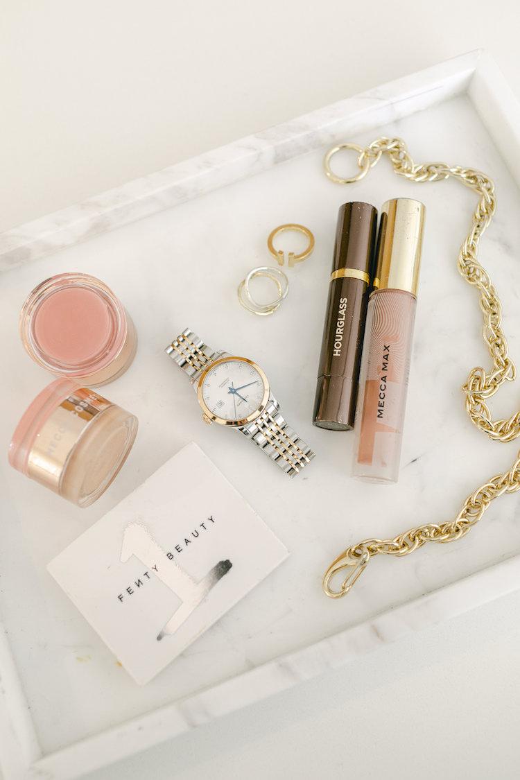 Marble tray with Fenty Beauty, Hourglass mascara, Mecca Max lip gloss, a watch, rings and gold chain