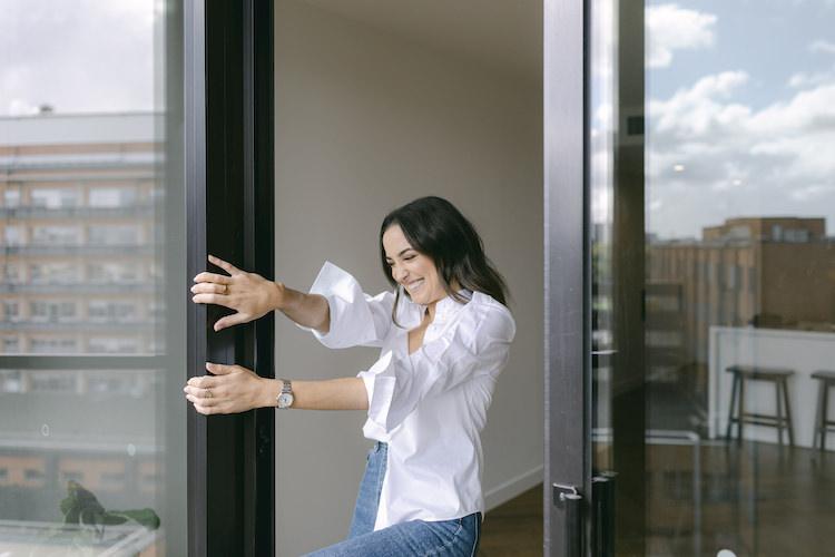 Abbey Gelmi laughing at a glass balcony door in a white shirt and jeans, city rooftop view