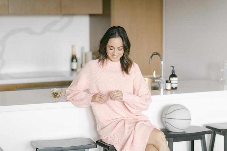 Abbey Gelmi in a pink knit sweater dress at a kitchen counter with champagne and a basketball nearby
