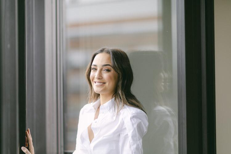Abbey Gelmi smiling through a glass door in a white shirt, her reflection visible, soft natural light