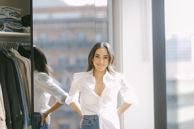 Abbey Gelmi standing by her wardrobe mirror in a white shirt and jeans, smiling, city view behind