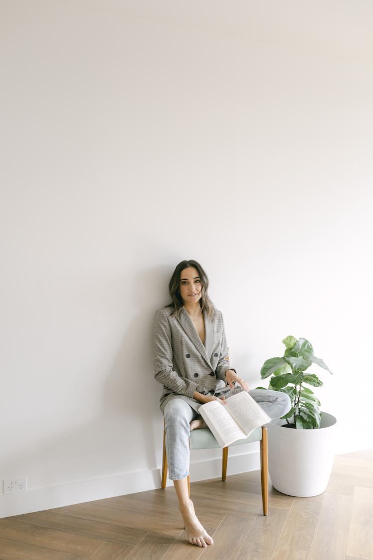 Abbey Gelmi barefoot on a chair with an open book, check blazer and jeans, fiddle-leaf fig beside her