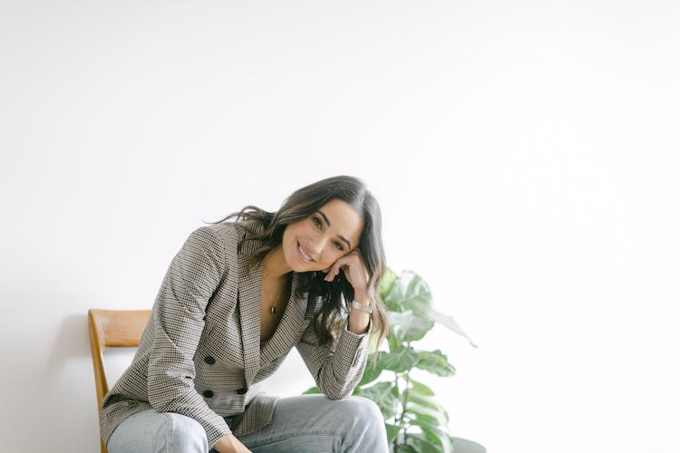 Abbey Gelmi leaning on her hand smiling, seated in a check blazer, fiddle-leaf fig in background