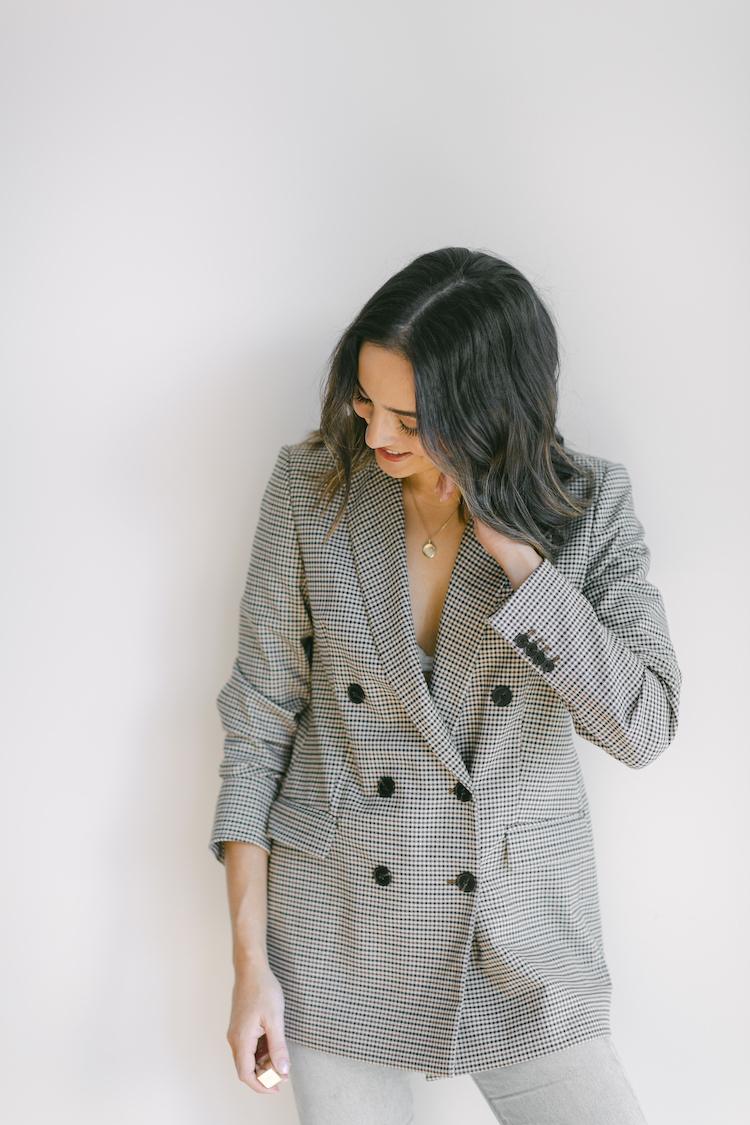 Abbey Gelmi looking down in an oversized houndstooth blazer with pendant necklace, white background