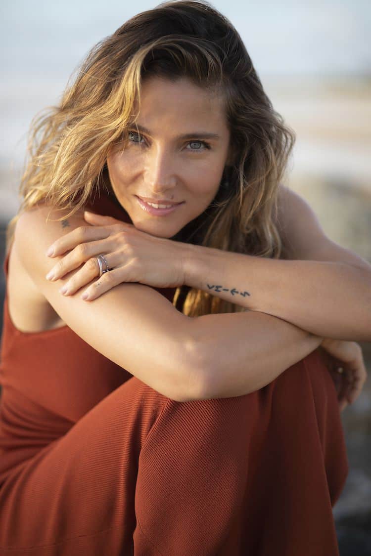 Elsa Pataky in a rust dress, arms crossed over her knees, smiling softly on the beach at golden hour