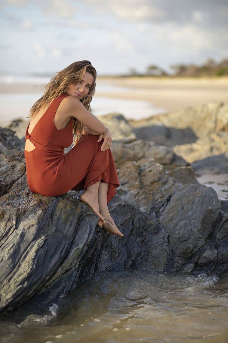 Elsa Pataky standing on a beach in a white flowing blouse, smiling with wind-swept hair at sunset