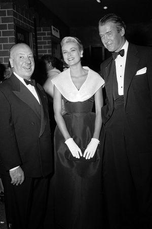 Grace Kelly in a dark gown with white collar standing between Alfred Hitchcock and a man in black tie
