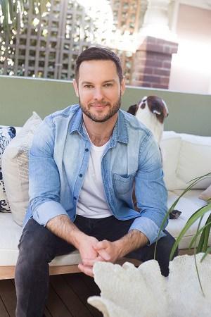 Darren Palmer seated on an outdoor sofa in a denim shirt, his dog peeking out from behind the cushions
