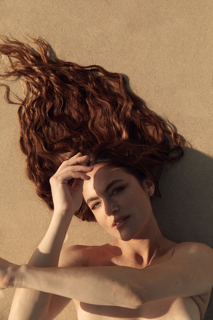 Julia Stone laughing on a rocky beach in a flowing white tulle gown, wind in her wavy hair at golden hour