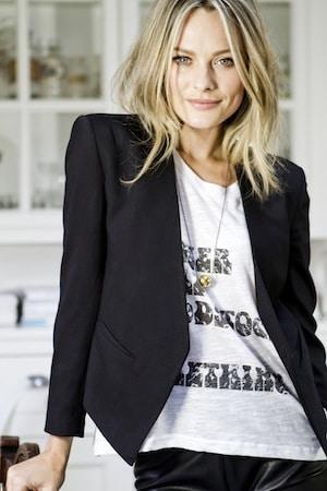 Sarah Jagger in a black blazer over a graphic tee with pendant necklace, tousled blonde hair, white kitchen behind