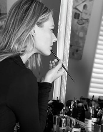Black-and-white profile of Sarah applying makeup in a mirror, hair pulled back, vanity with brushes visible