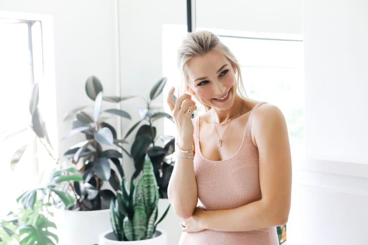 Anna Heinrich smiling with one hand raised in a blush tank top, monstera and fiddle-leaf fig behind