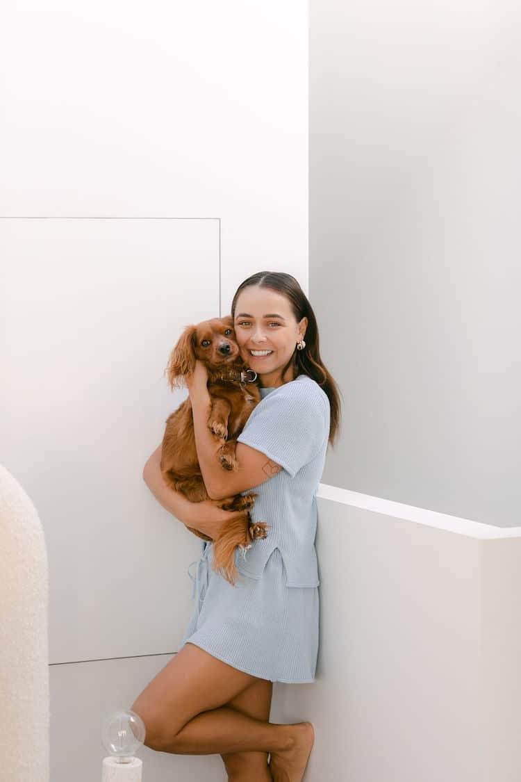 Brooke Blurton in a pale blue tee cuddling her ruby Cavalier King Charles Spaniel against a white wall