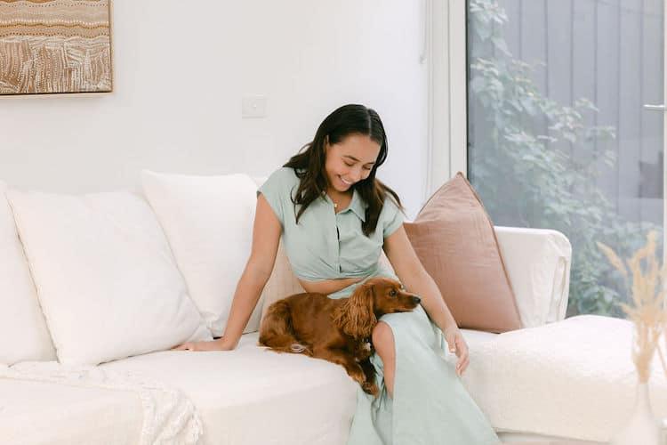 Brooke Blurton on a cream couch smiling down at her dachshund in a sage green ribbed top, soft natural light