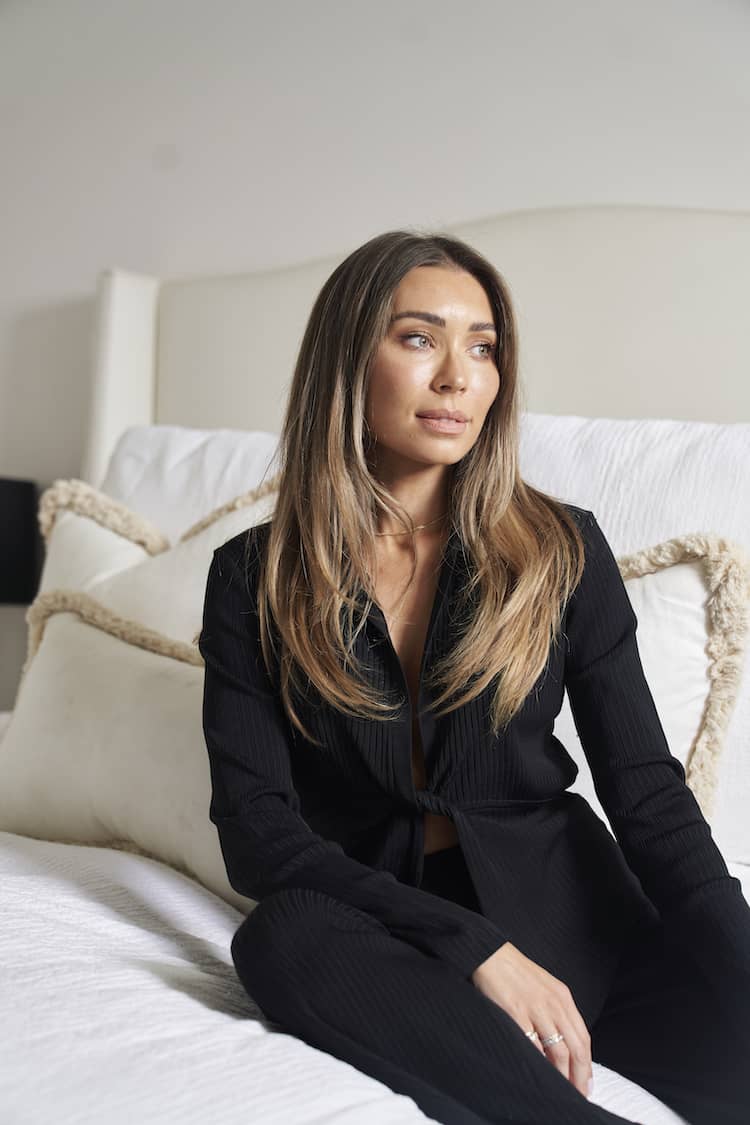 Rozalia seated on white bed in black ribbed suit, hair down, gazing left with natural makeup