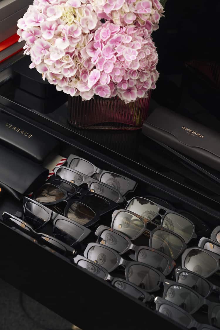 Designer sunglasses collection in drawer with Versace and Oliver Peoples cases, pink hydrangeas above