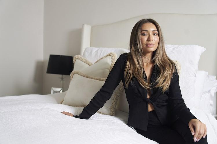 Rozalia Russian seated on her bed in a black knotted top with white linen and tasselled cushions
