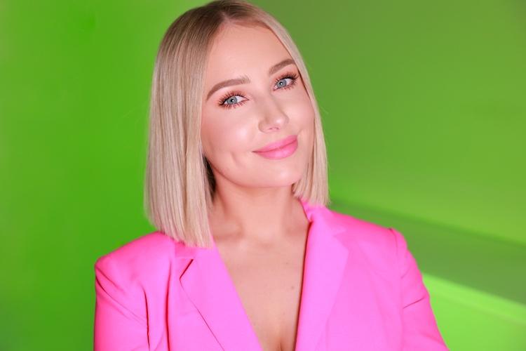 Lauren Curtis with a blonde bob and pink blazer, glowing pink makeup against a green backdrop