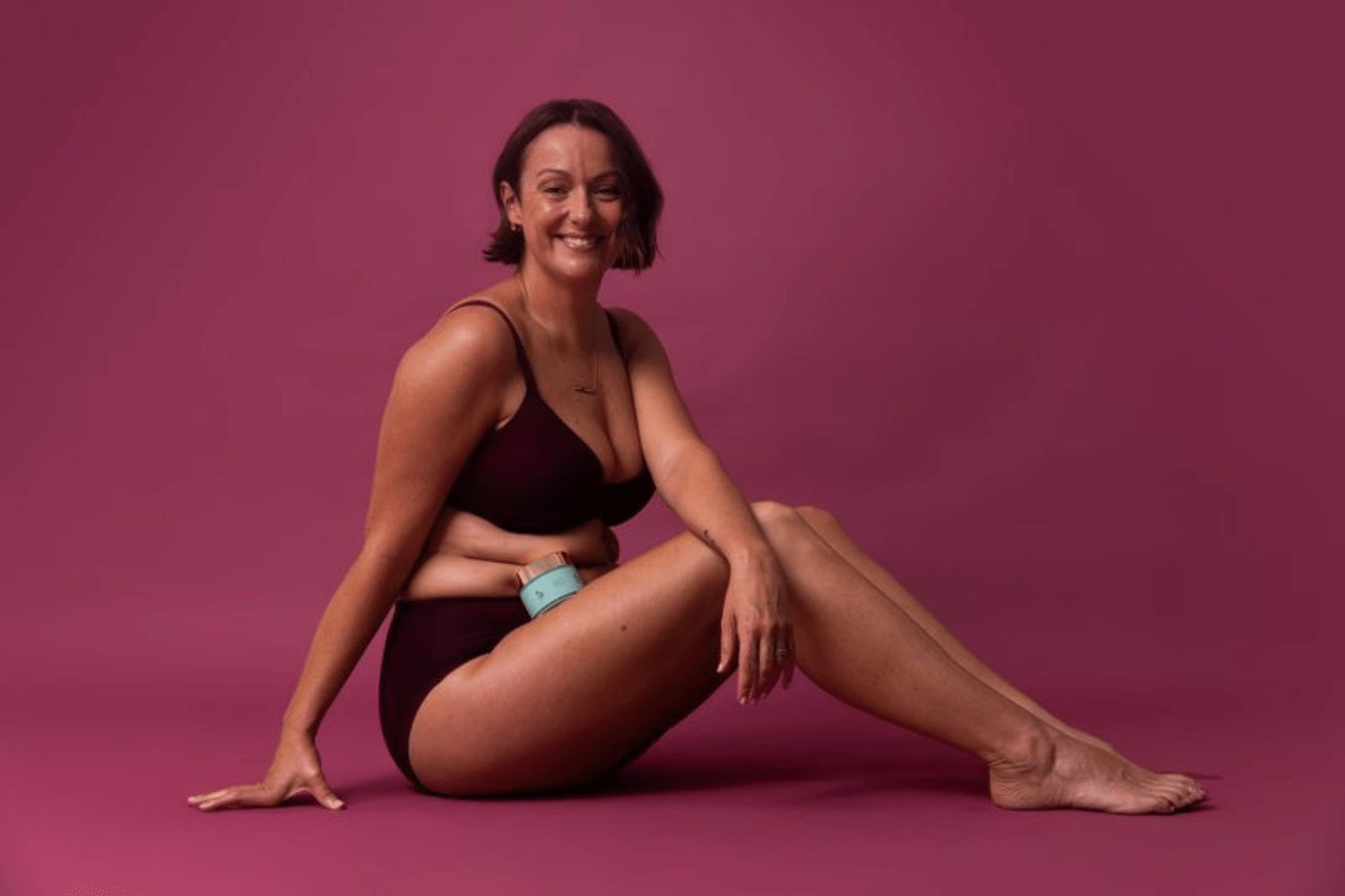 Celeste Barber sitting on the floor in burgundy underwear, smiling, holding her Booie body cream