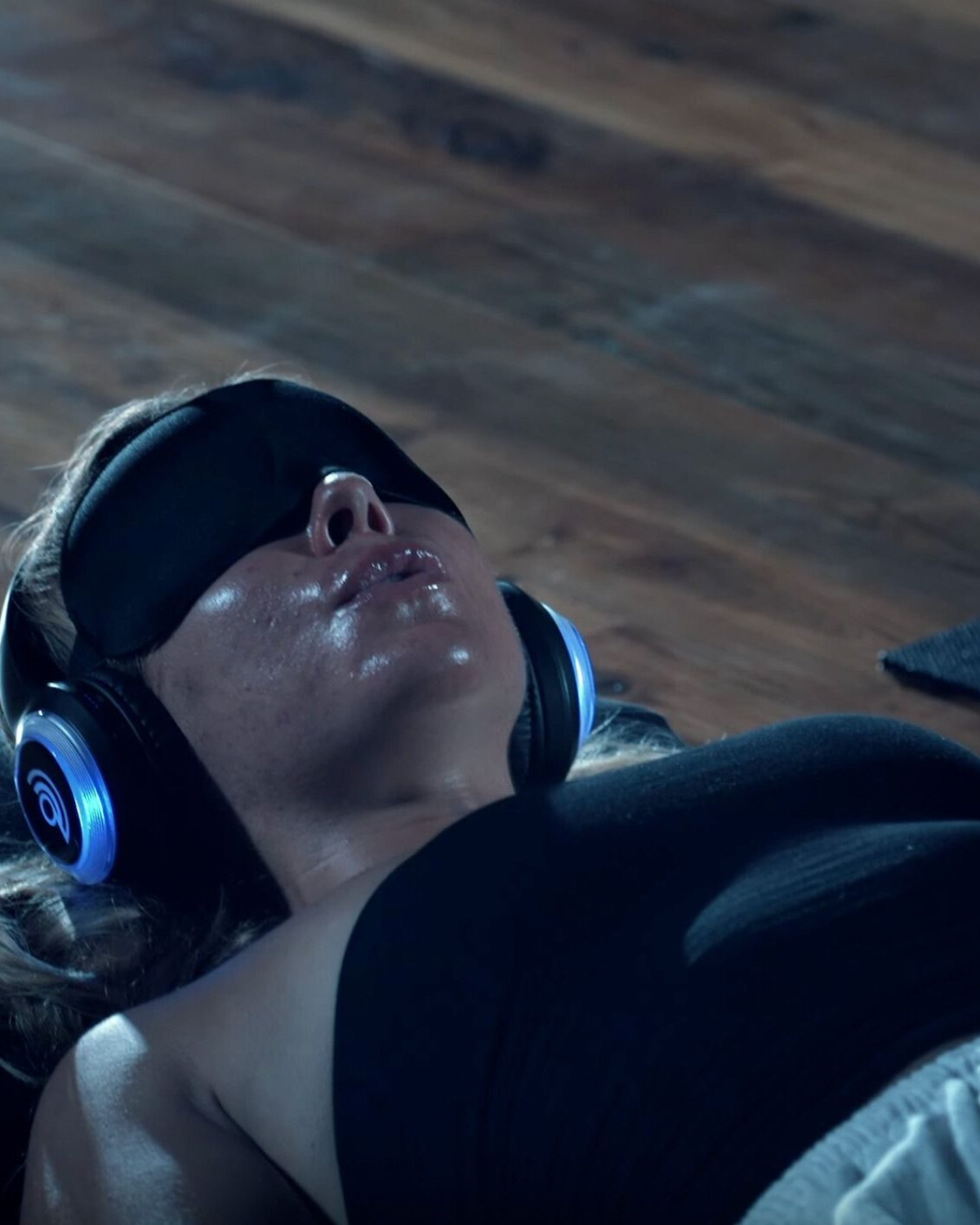 Close-up of a woman lying on a wooden floor during a 9D breathwork session, wearing noise-cancelling headphones with blue LED rings and a black eye mask, her face glistening with perspiration