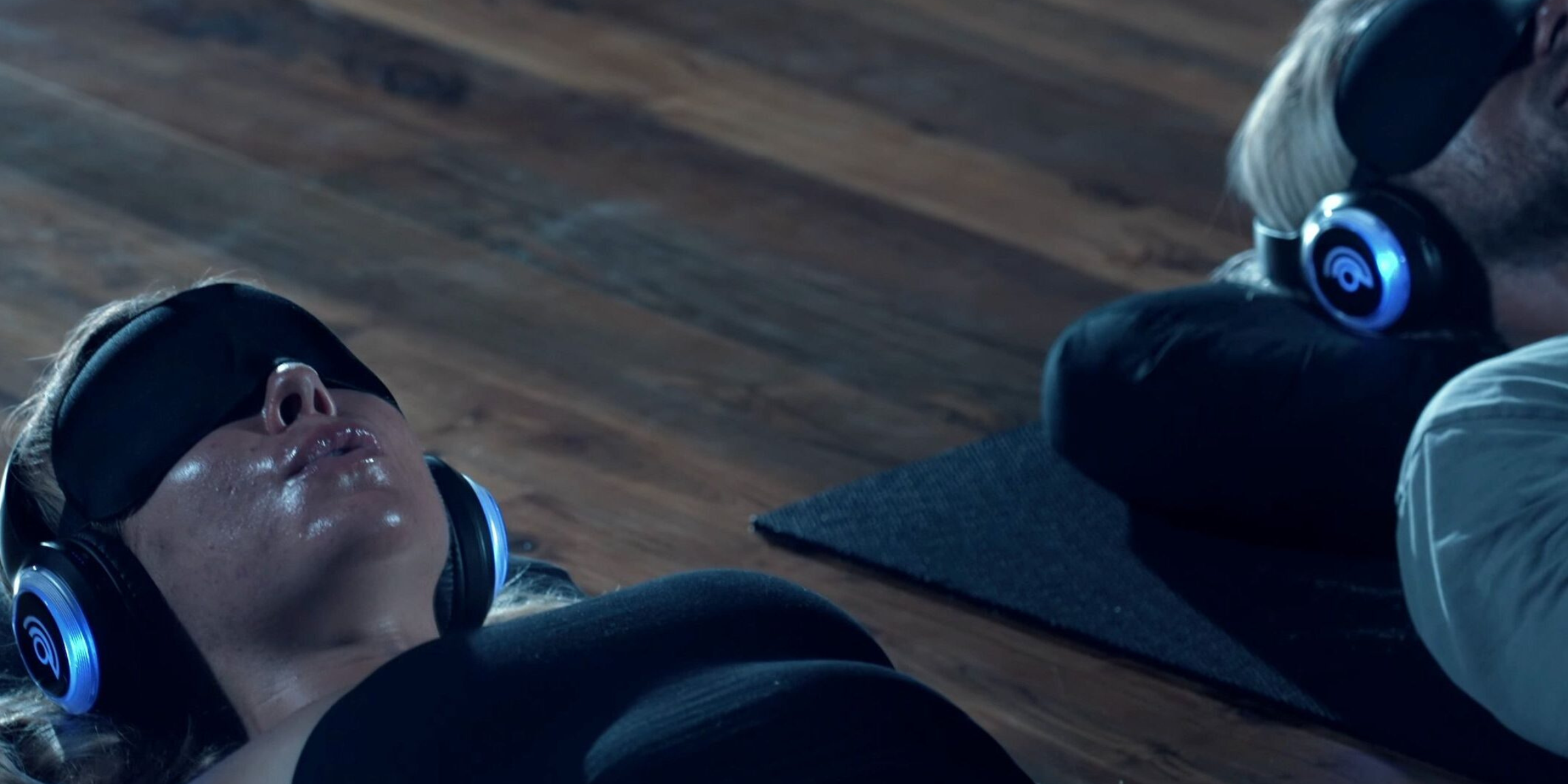 A group of people lying on yoga mats during a 9D breathwork session in a dim studio, wearing headphones and eye masks, with soft ambient lighting